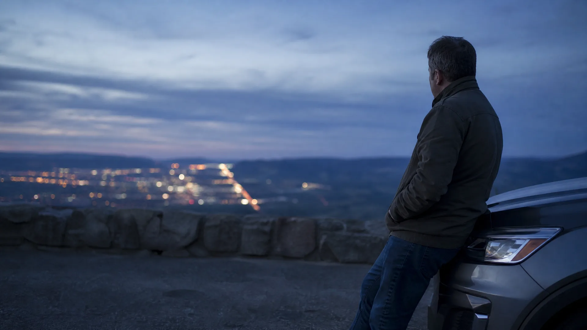 A man in a dark jacket and jeans leans against the front of an SUV at a stone-walled overlook at dusk, facing distant city lights in a valley.